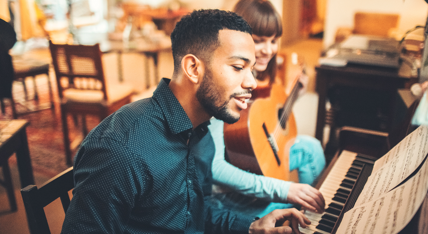 Person playing an upright piano with sheet music, accompanied by guitarist in a cozy room.